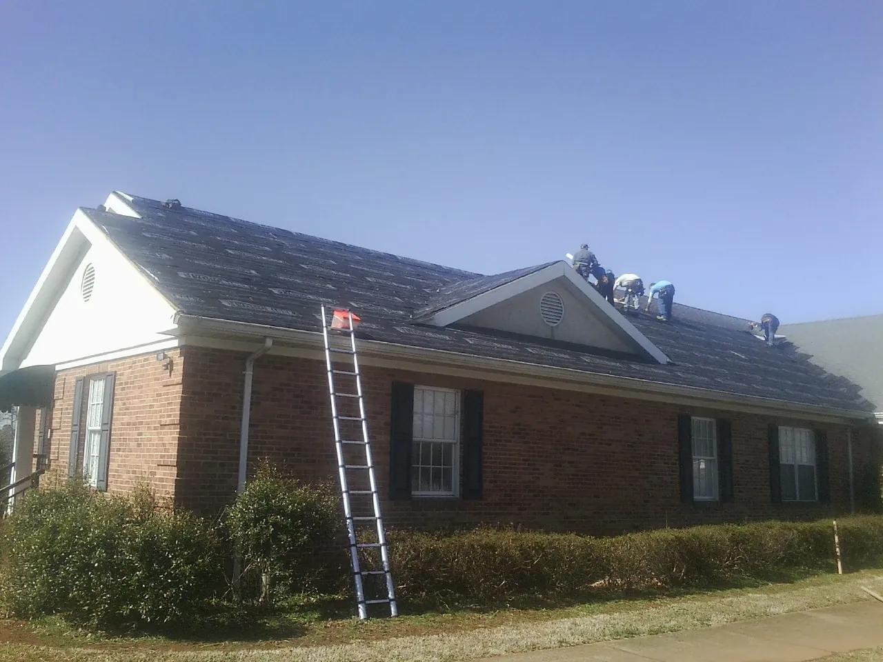 Crew tearing shingles off a brick ranch roof with a ladder set — Emergency Roof Repair work by our crew in Bunker Hill